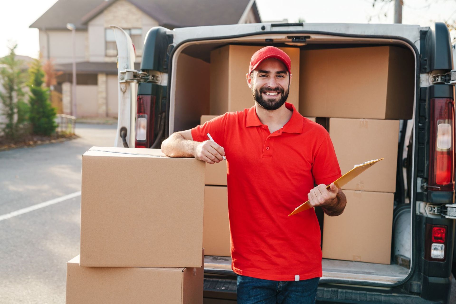 Delivery worker in red shirt and cap unloading cardboard boxes from a van, smiling outdoors