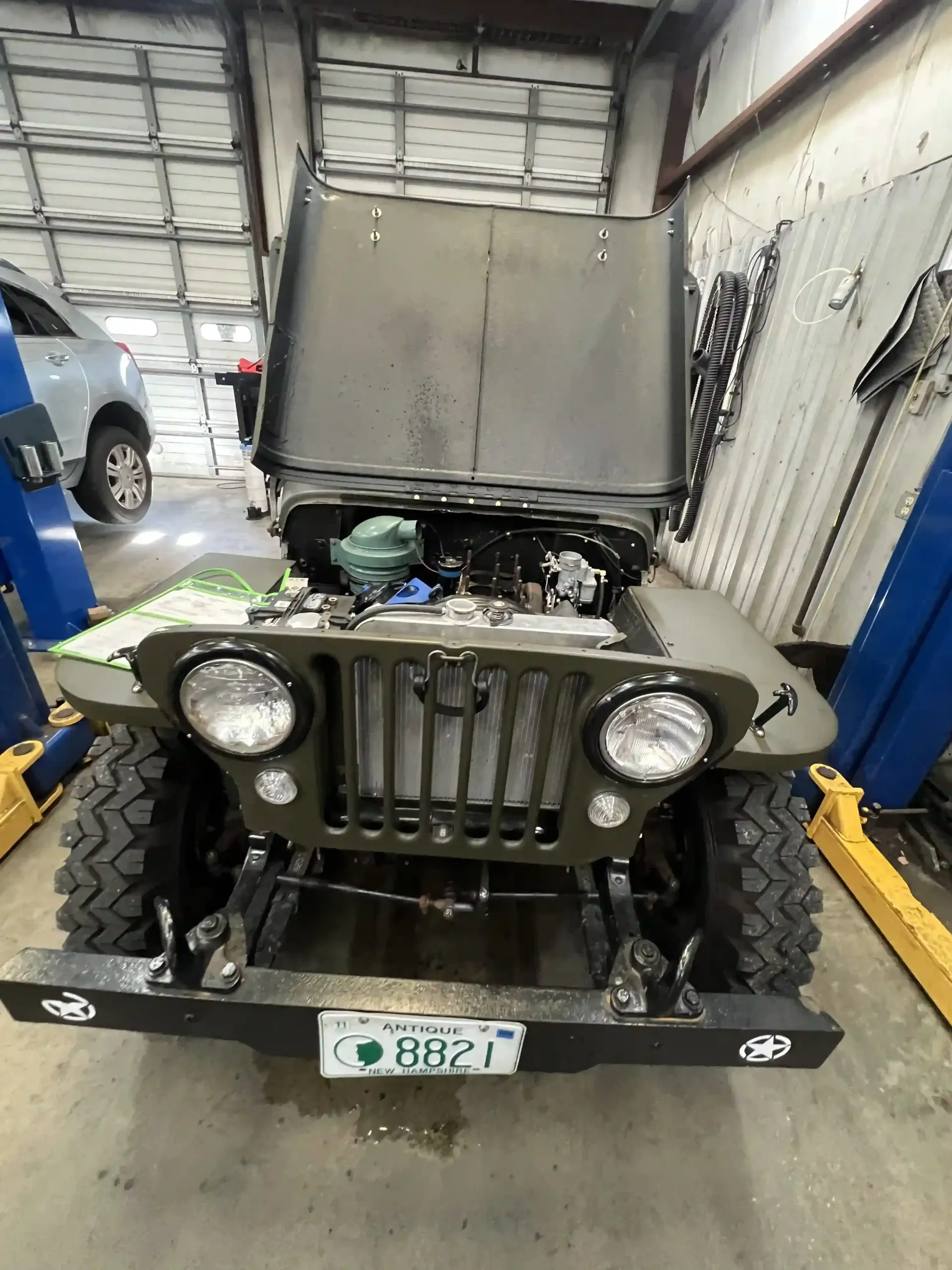 Front view of a vintage olive-green Jeep with hood open in a garage, showing the engine area.