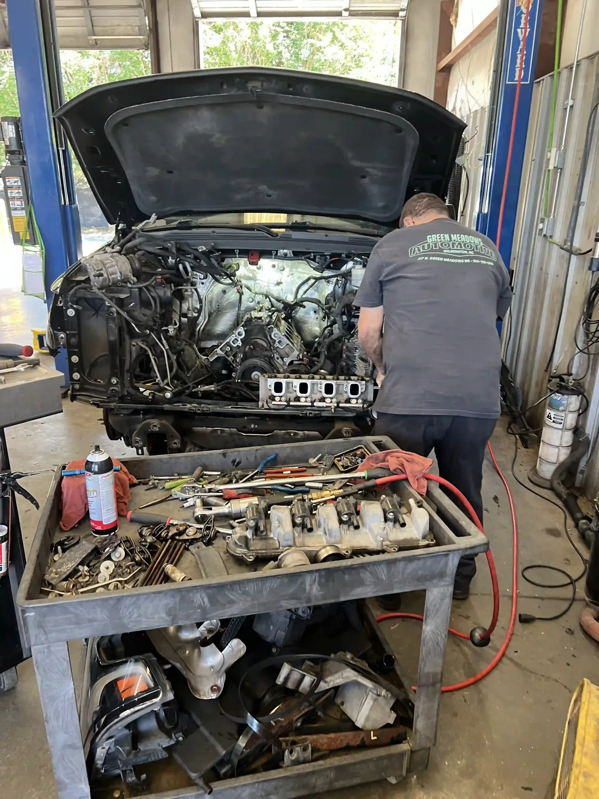 Mechanic working on a car with hood open in a garage, tools on a cart in front.