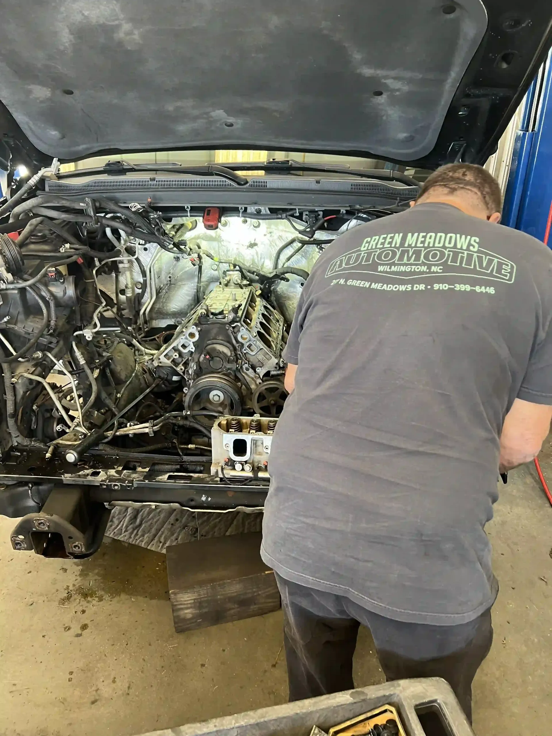 Mechanic working on a car with the hood open in a garage, engine bay exposed and damaged.