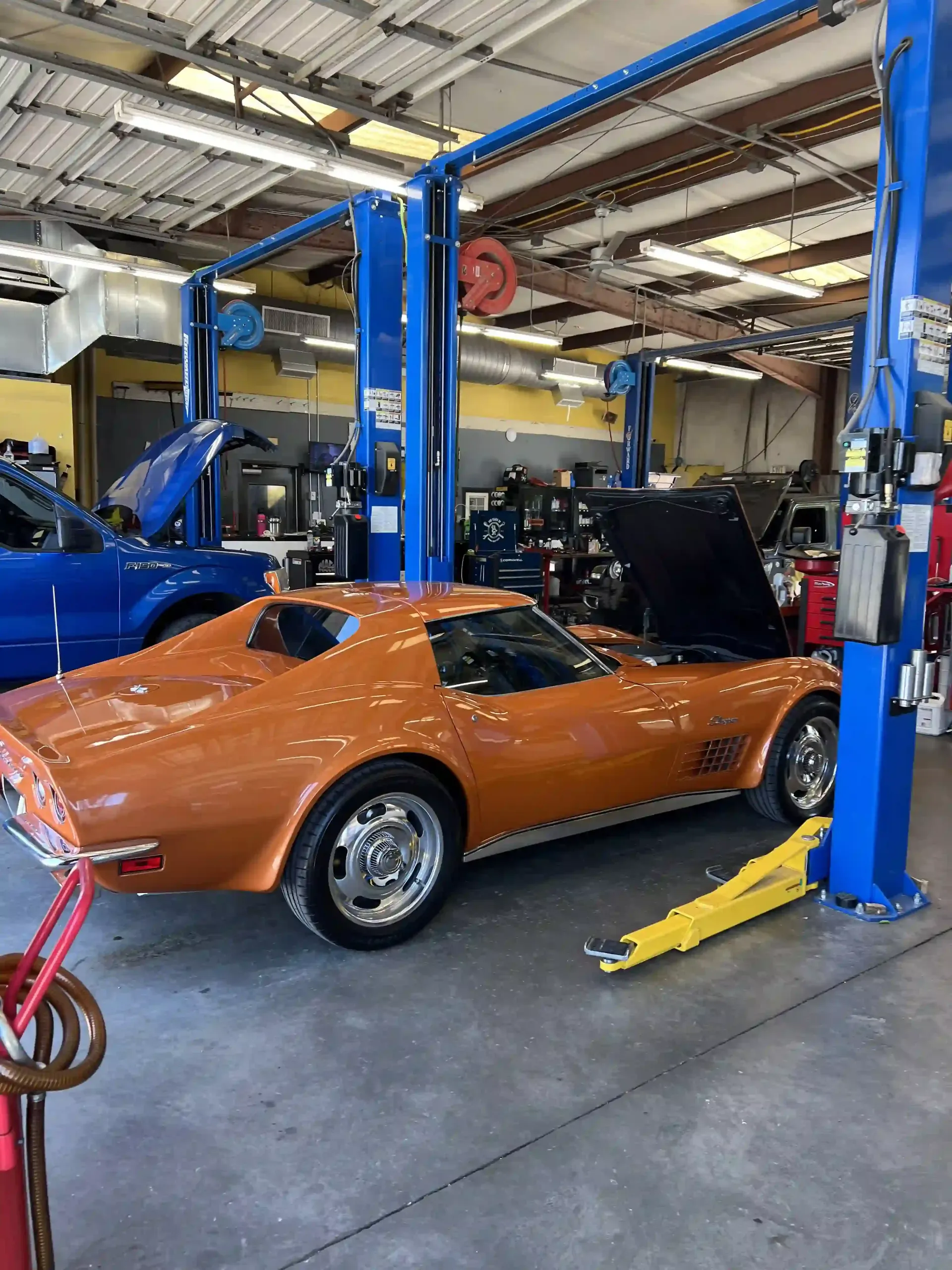 Orange classic car in an auto repair shop on a lift with blue equipment and open hood
