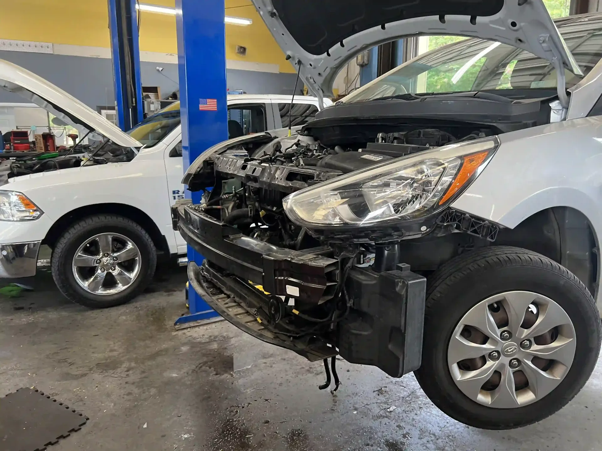 Two cars in an auto repair shop, one with its front end damaged and hood open.