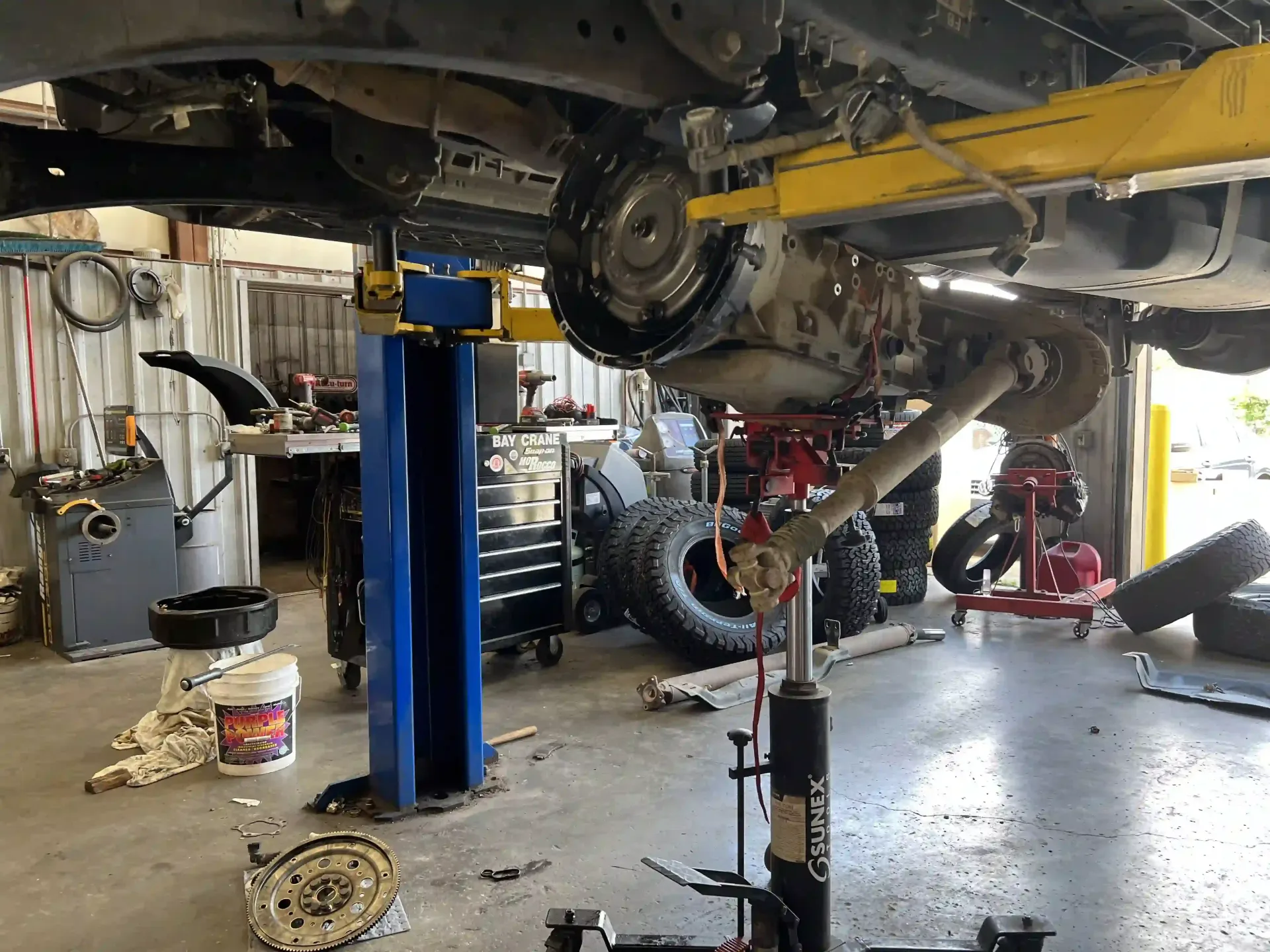 Car lifted on a blue hydraulic jack in an auto repair shop, with tools and wheels on the floor.