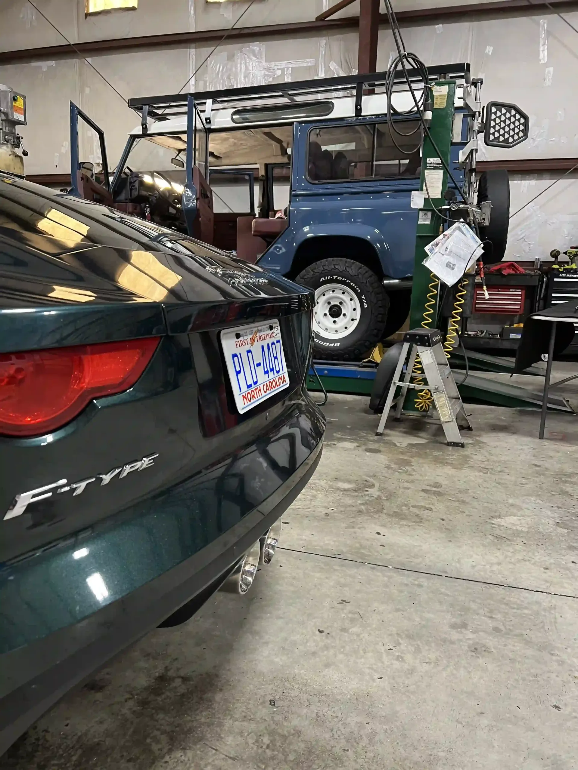 Dark green car in a garage beside a blue off-road truck on a lift with a ladder nearby