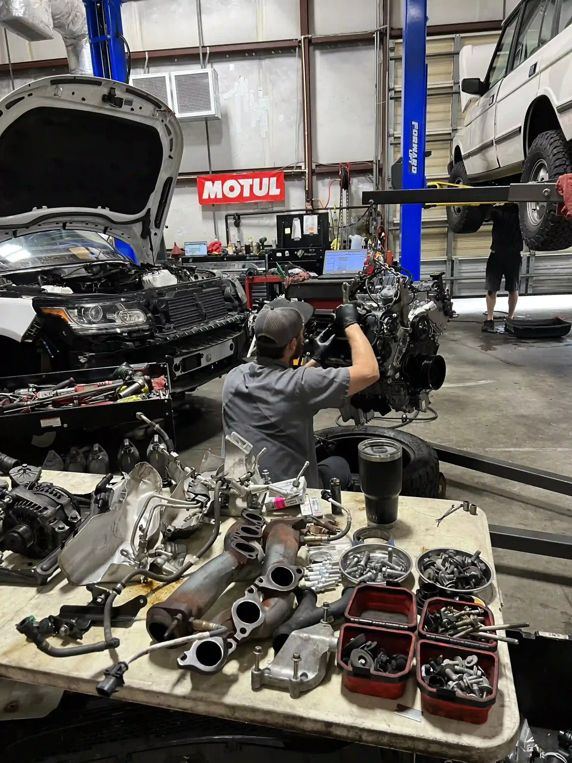 Auto repair shop with mechanic working on a car engine amid disassembled parts and tools.