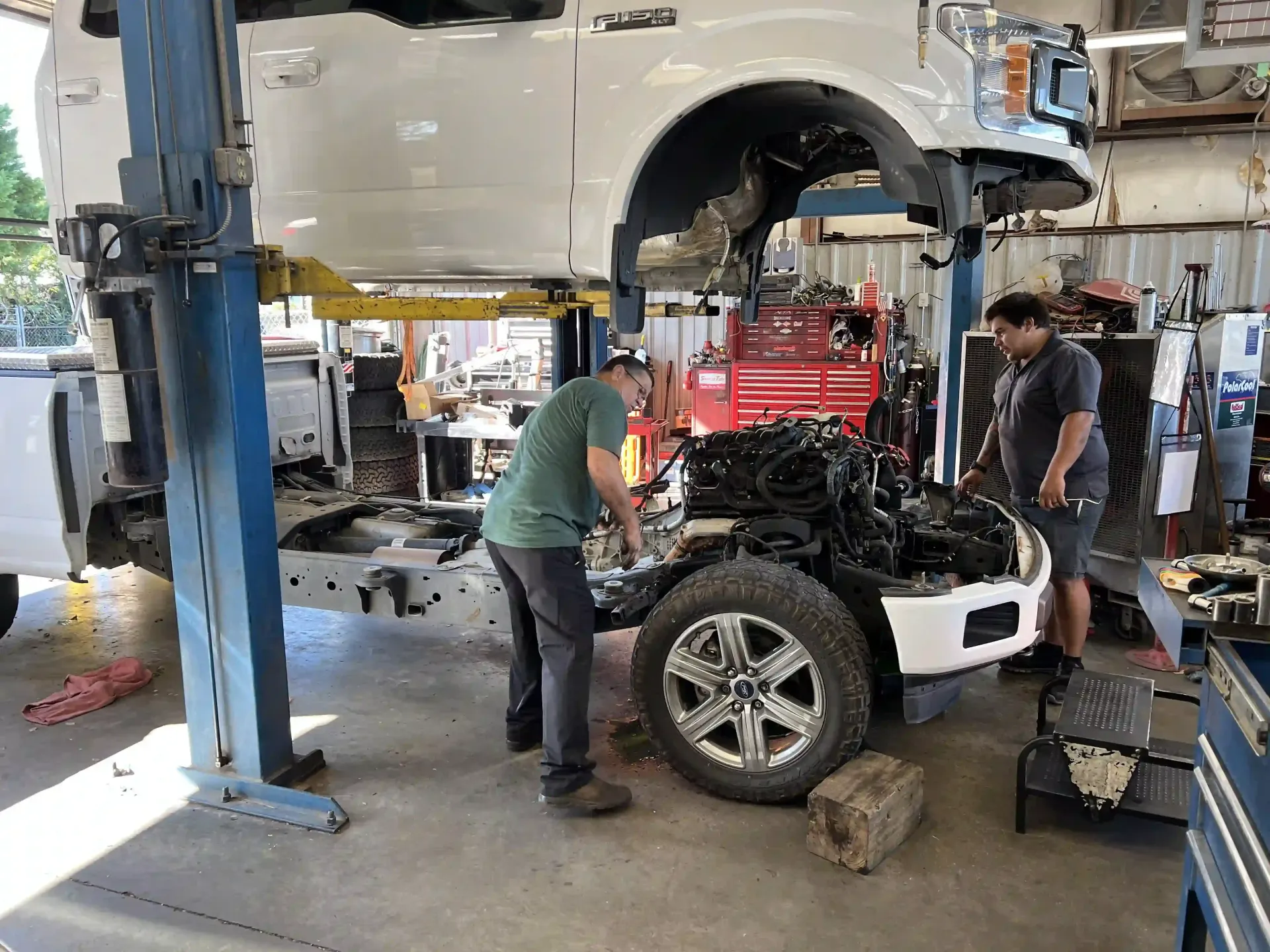 Mechanic works on a white SUV in a busy auto shop, with another vehicle lifted overhead.