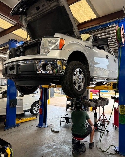 Silver pickup truck lifted on a shop hoist while a mechanic works underneath in an auto garage.