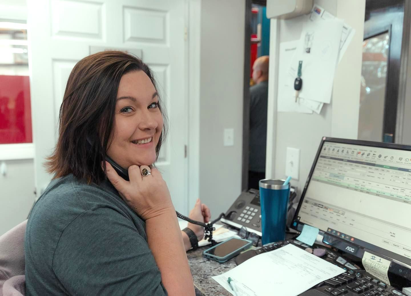Woman smiling at a desk with dual monitors, paperwork, and a blue tumbler in an office.