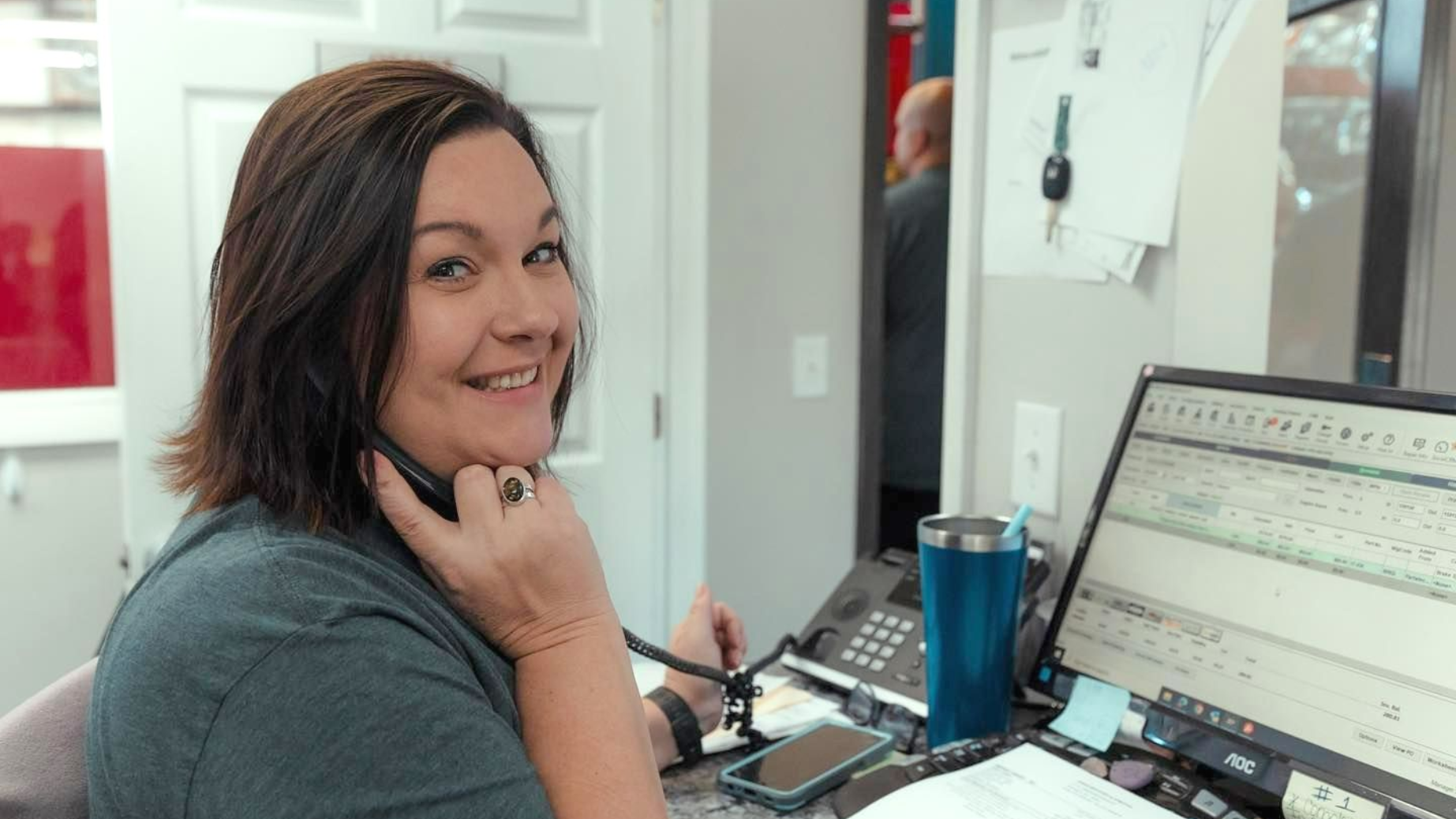 Woman smiling at a desk with a computer monitor, holding a phone in an office setting
