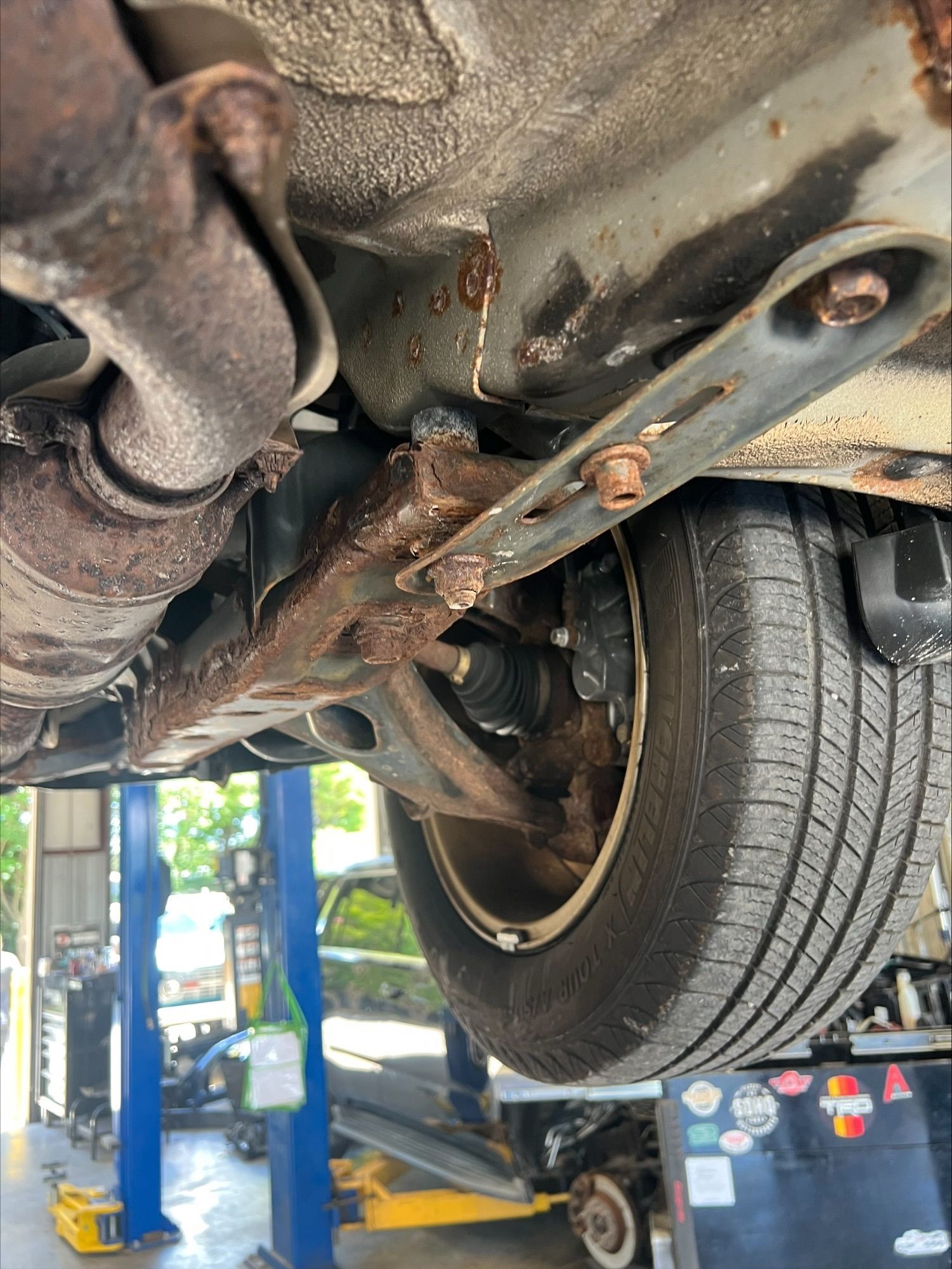 Undercarriage of a car on a lift in an auto repair shop, with a wheel and suspension visible.