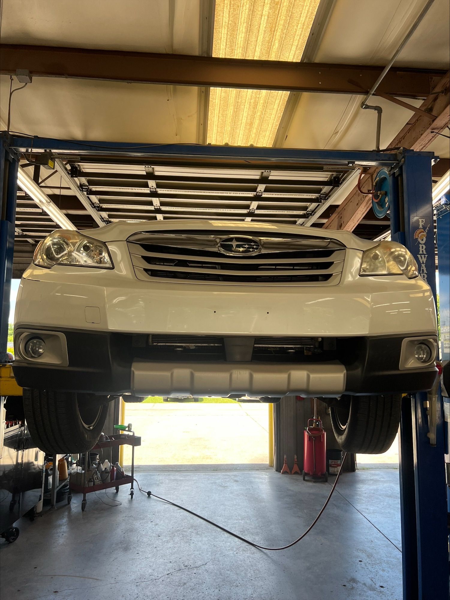 White SUV raised on a lift in an auto repair garage, viewed from below with hood open.
