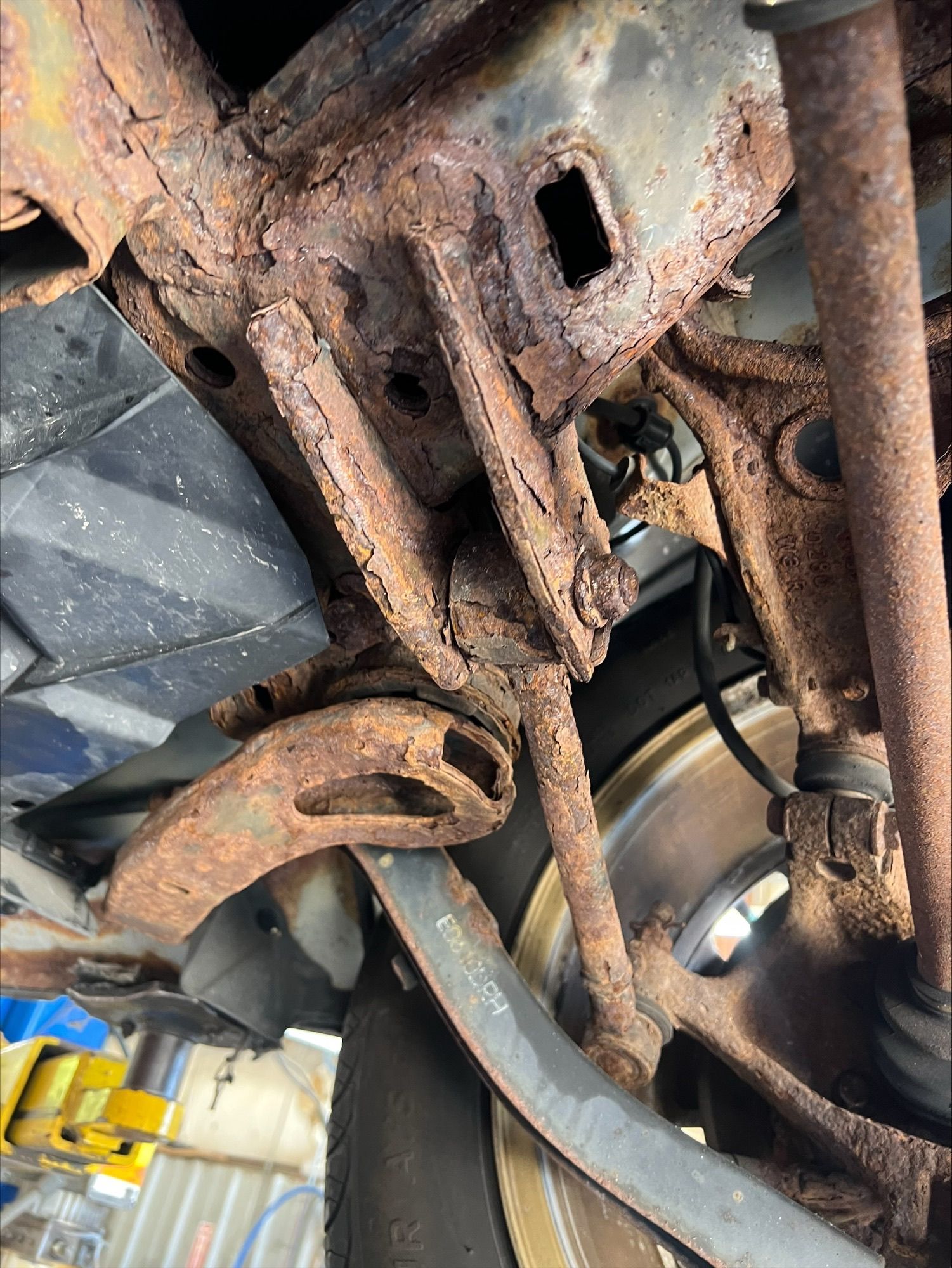 Rusty undercarriage and suspension components beneath a vehicle, with a wheel and brake assembly visible
