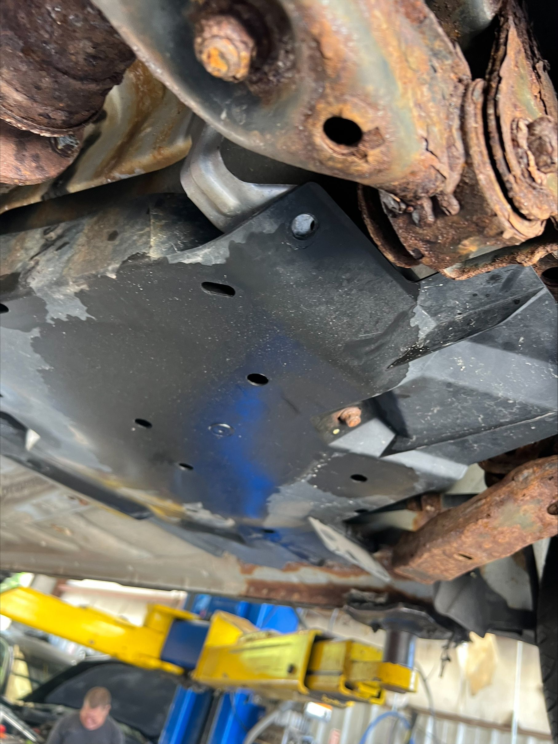 Undercarriage of a vehicle on a lift, showing rusted suspension parts and a black skid plate with blue stain