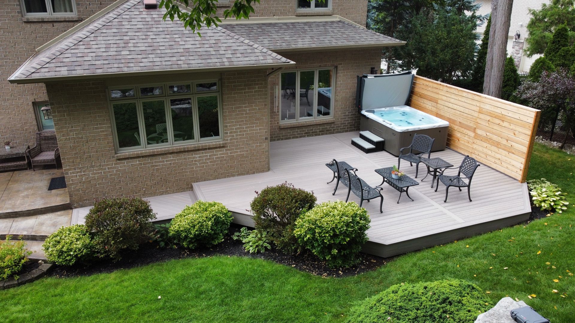 Back patio with outdoor dining table and chairs beside a brick house, surrounded by shrubs and lawn.