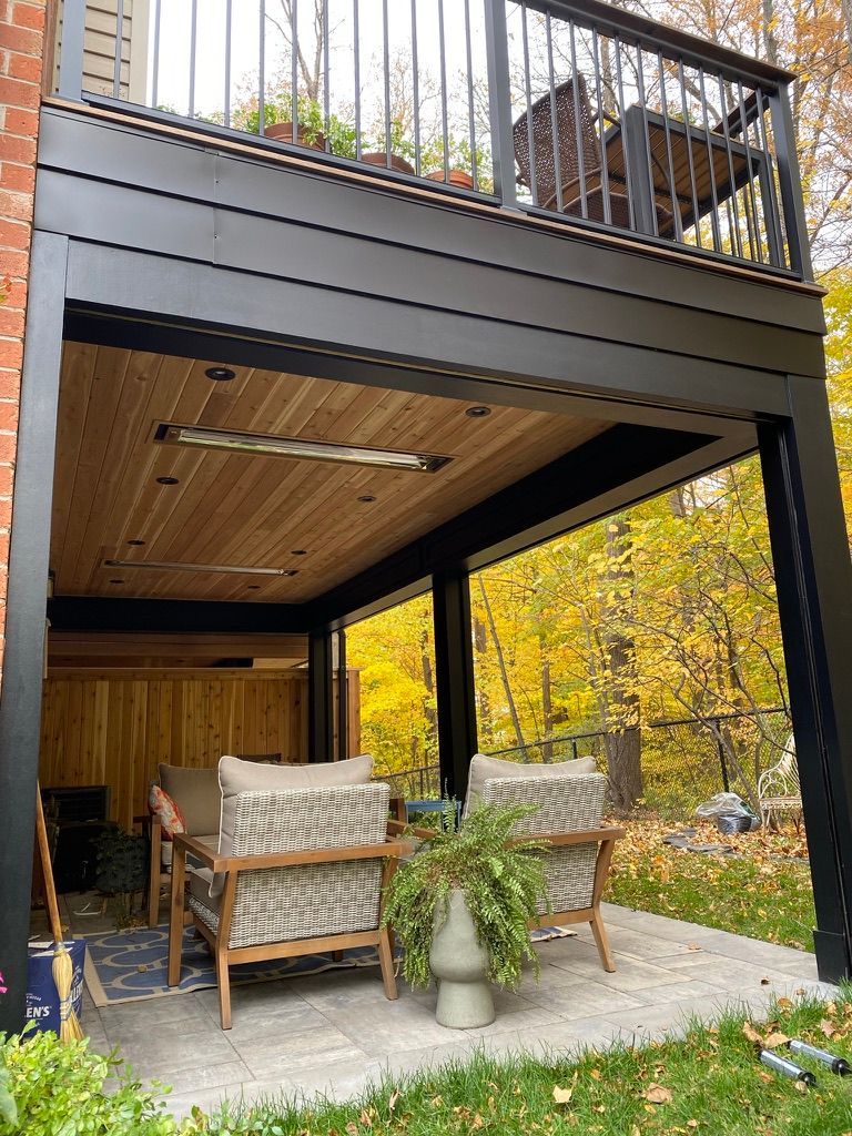 A cozy patio area under a wooden deck, featuring two armchairs, a potted plant, and a heater on the ceiling.