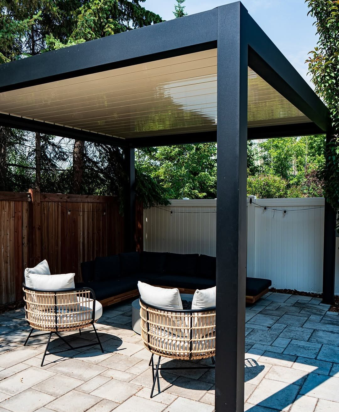 Modern backyard patio with a black pergola, wicker chairs, and a dark sectional sofa on stone pavers.