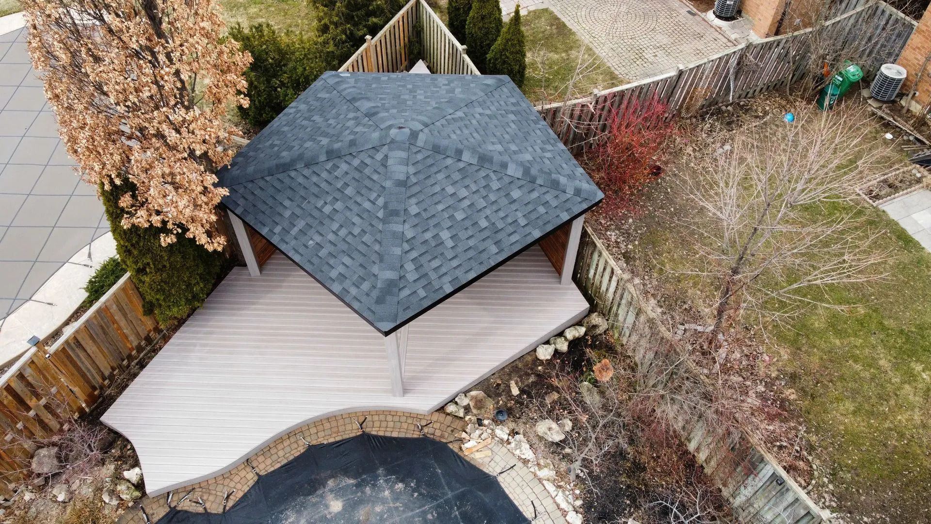 Aerial view of a backyard with a dark-roofed gazebo, wooden deck, pool edge, and fenced landscaping.