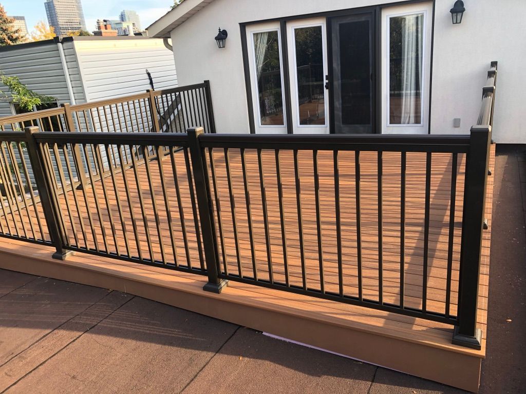 An elevated brown deck with black metal railing, featuring a glass door entryway to a white building.