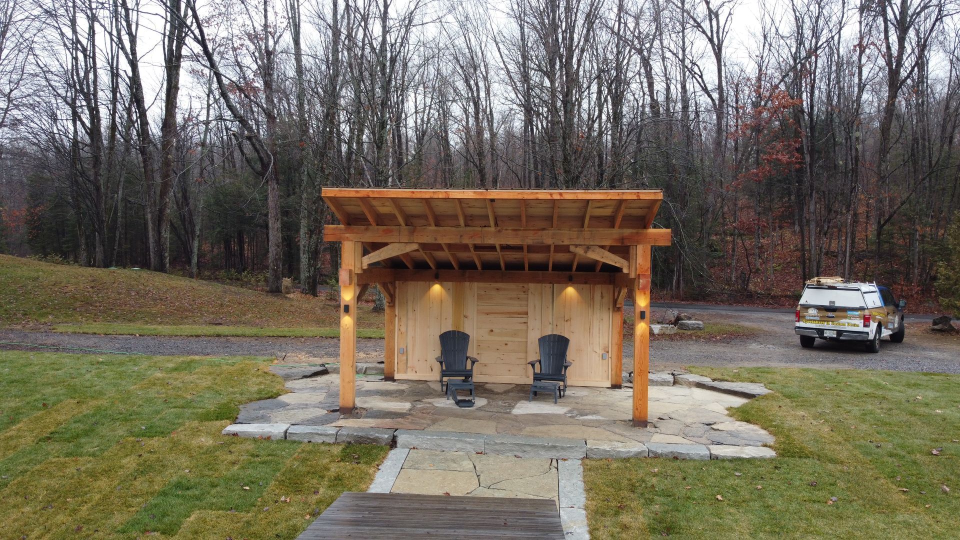Wooden shelter with two chairs on a paved pad in a wooded park, with a truck parked nearby.