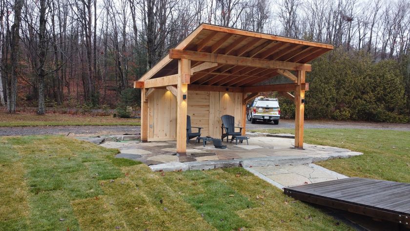 Wooden pavilion shelter with a grill and parked van beside a wet concrete pad in a grassy park.