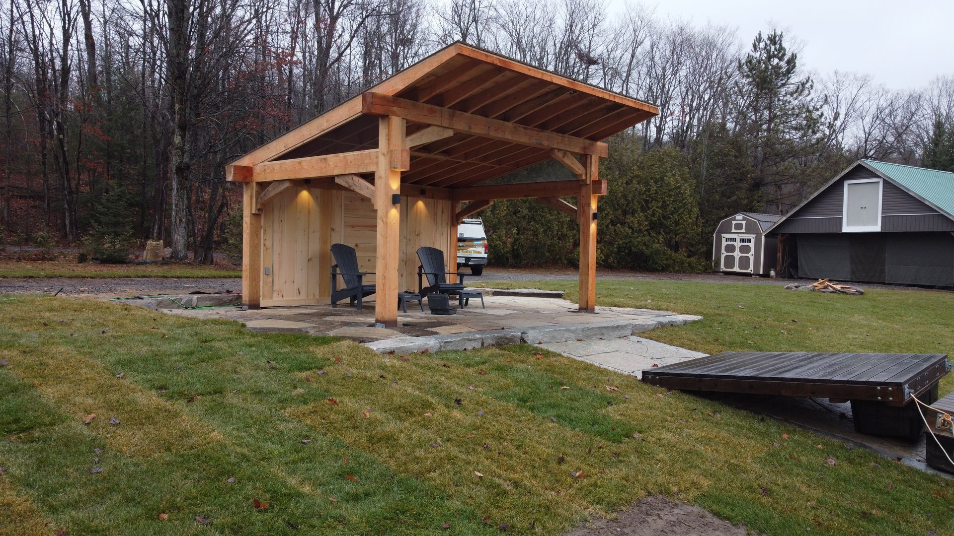 Wooden pavilion with a grill on a gravel yard beside a house, surrounded by trees in autumn.