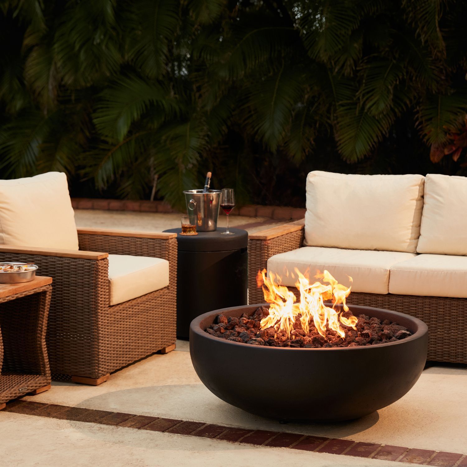 Outdoor patio with two wicker chairs and a round fire pit burning at dusk
