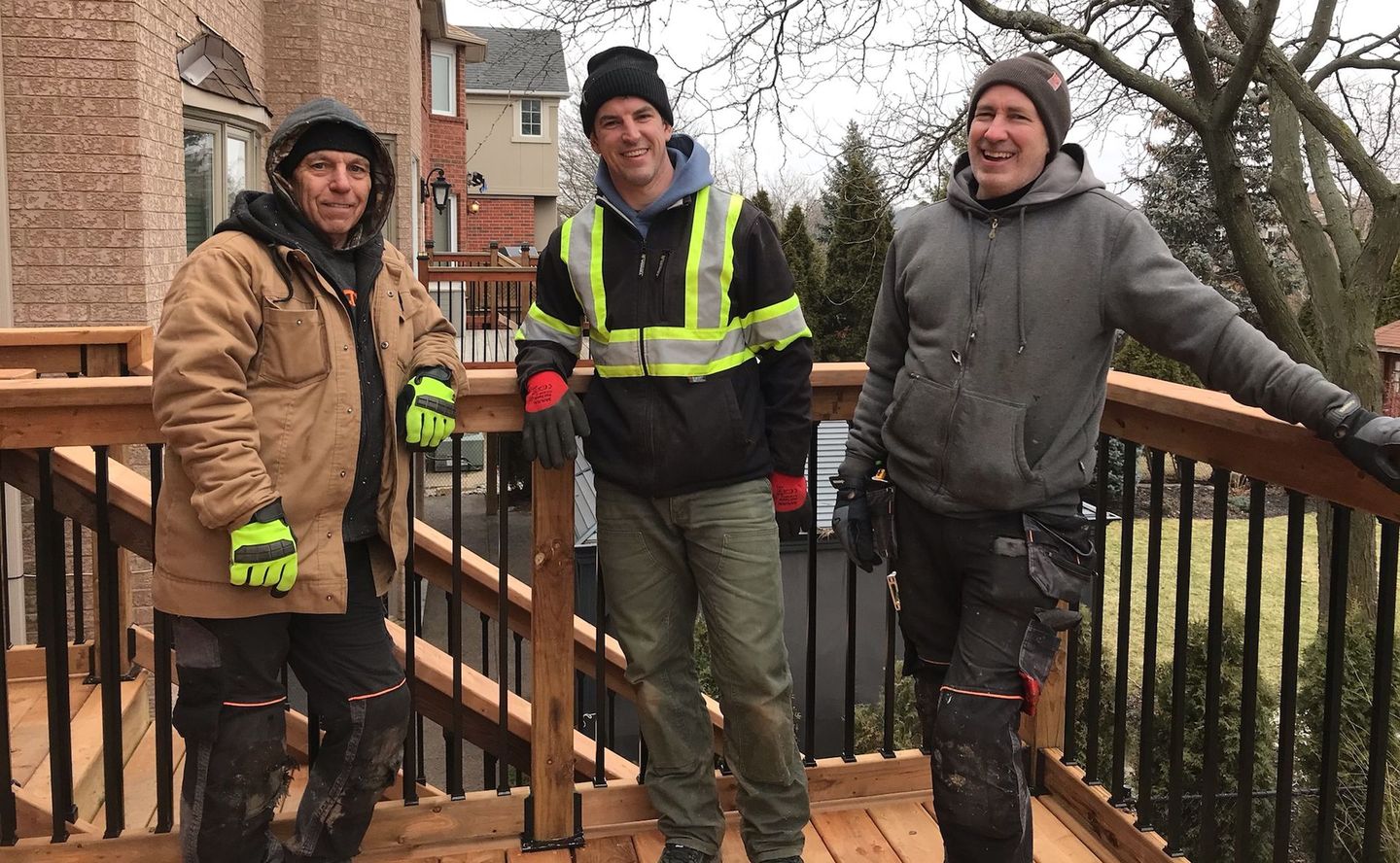 Three people in winter jackets standing on wooden steps by a railing outside a brick building.