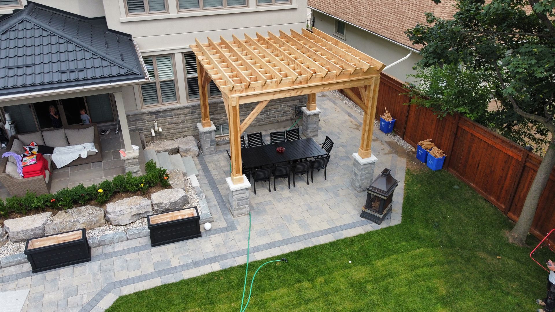 Wooden pergola over a dining table on a patio, adjacent to a house. Green grass and a wooden fence surround it.