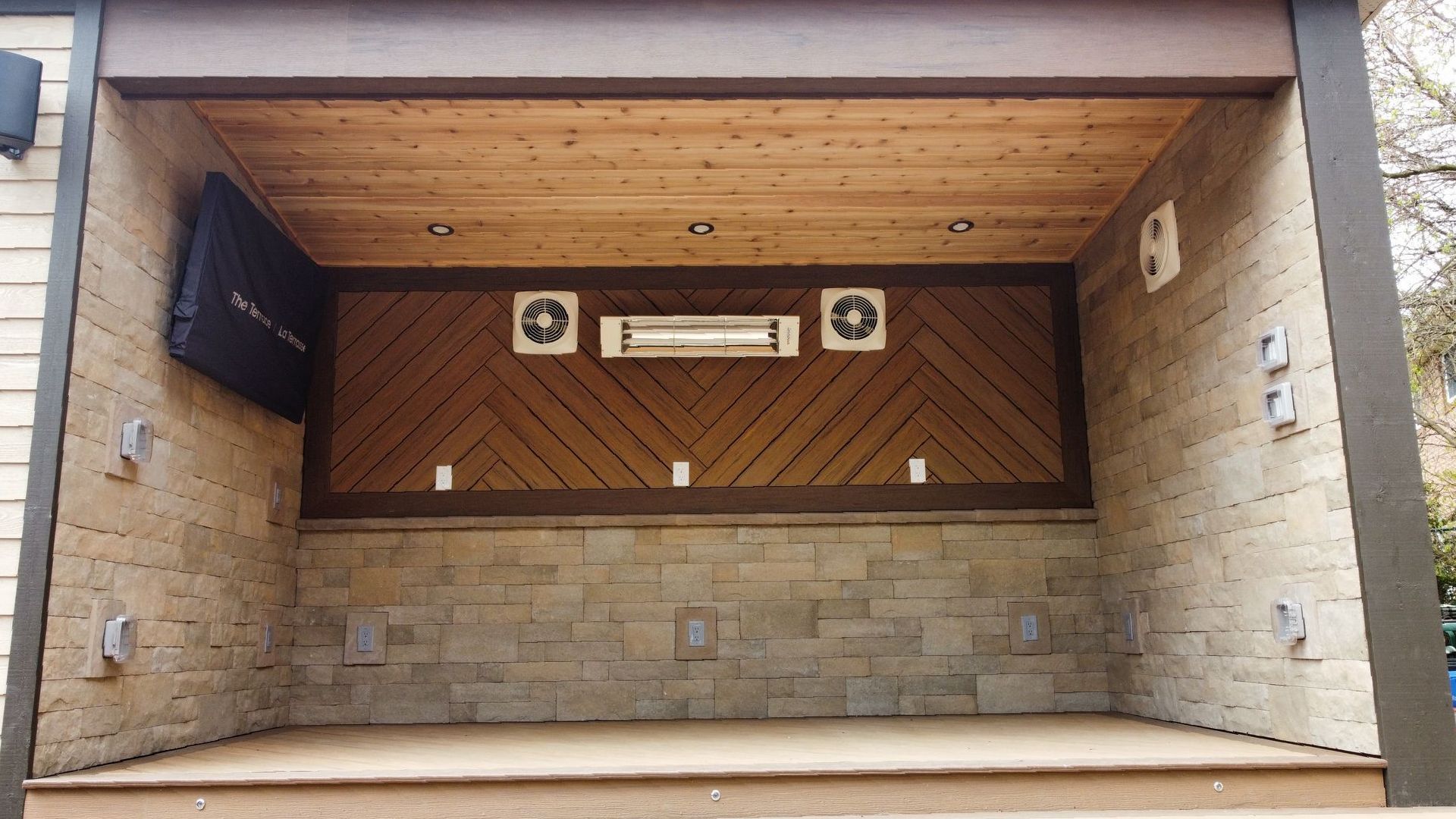 Covered outdoor space with wooden ceiling and accent wall, stone wall. Features a TV, two fans, and a heater.