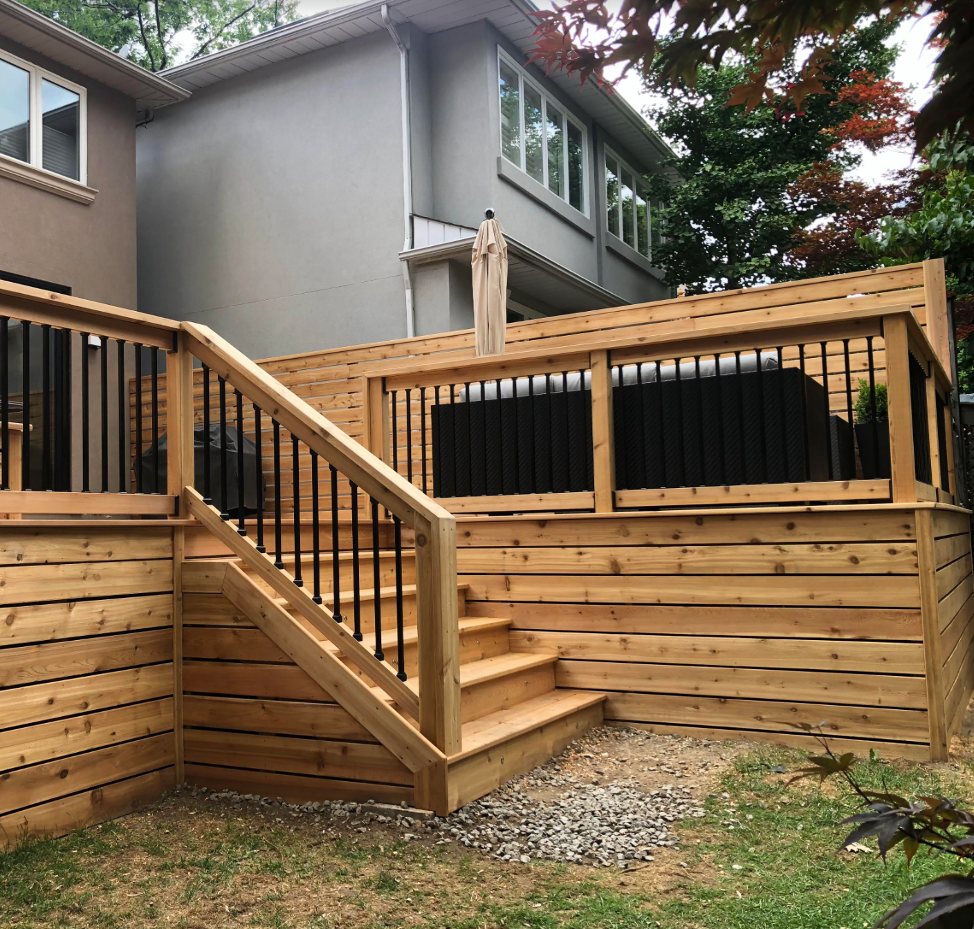 Wooden backyard deck with stairs and railing beside a gray house, surrounded by trees and gravel.