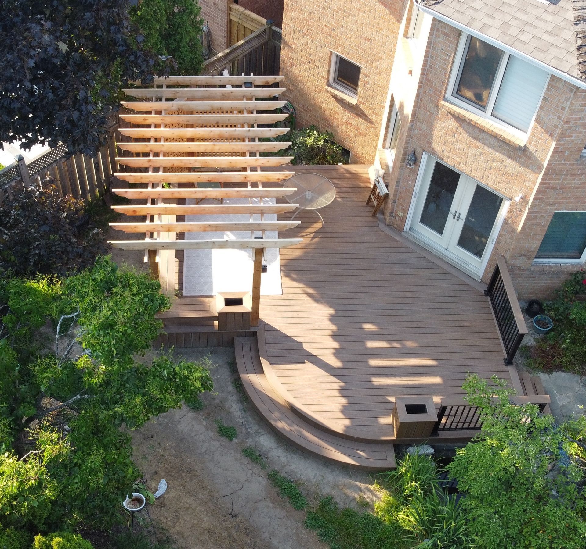 Aerial view of a backyard deck with pergola beside a brick house, surrounded by trees and garden path.
