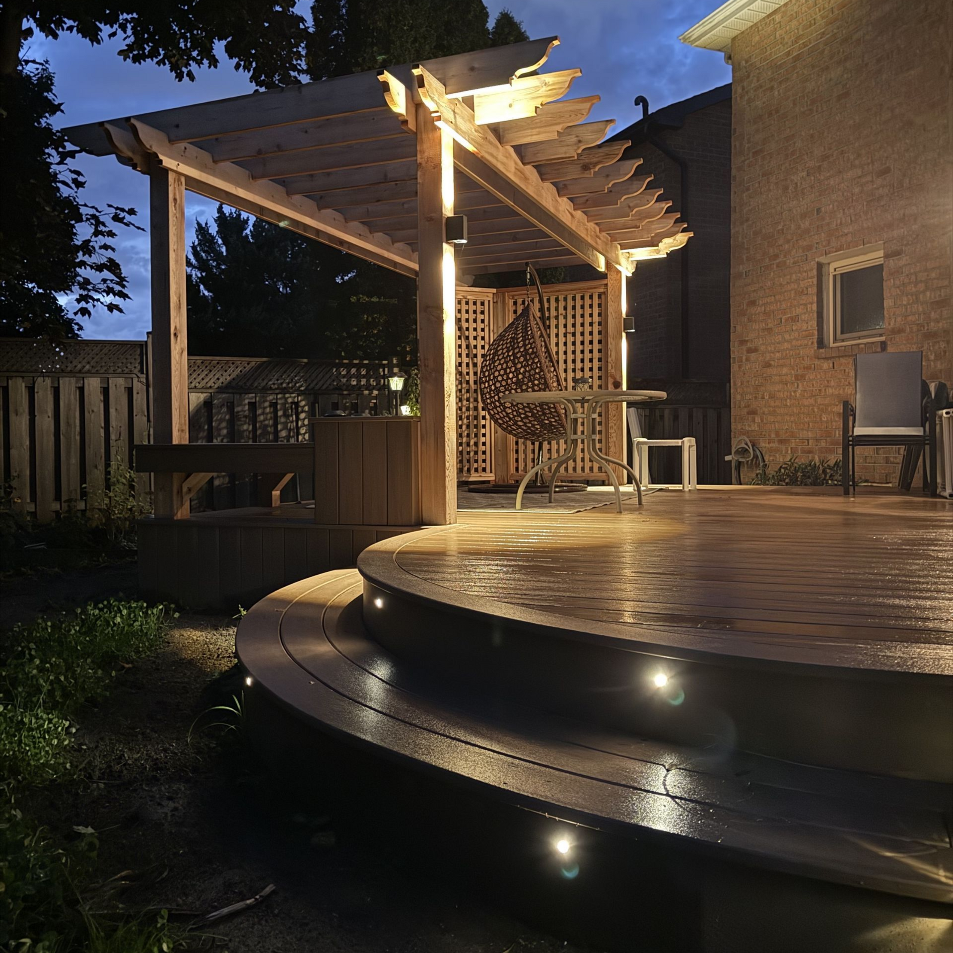 Warm-lit backyard deck with pergola at dusk, string lights, and curved steps along a patio.