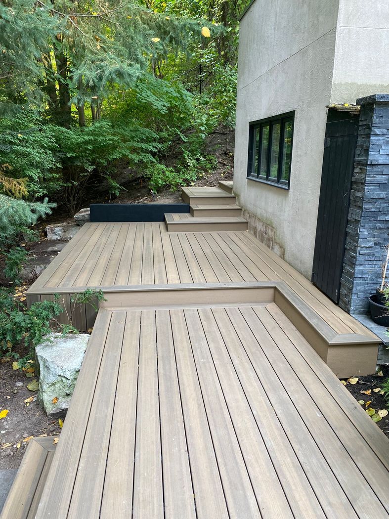 Wooden deck and steps beside a modern house, surrounded by dense greenery.