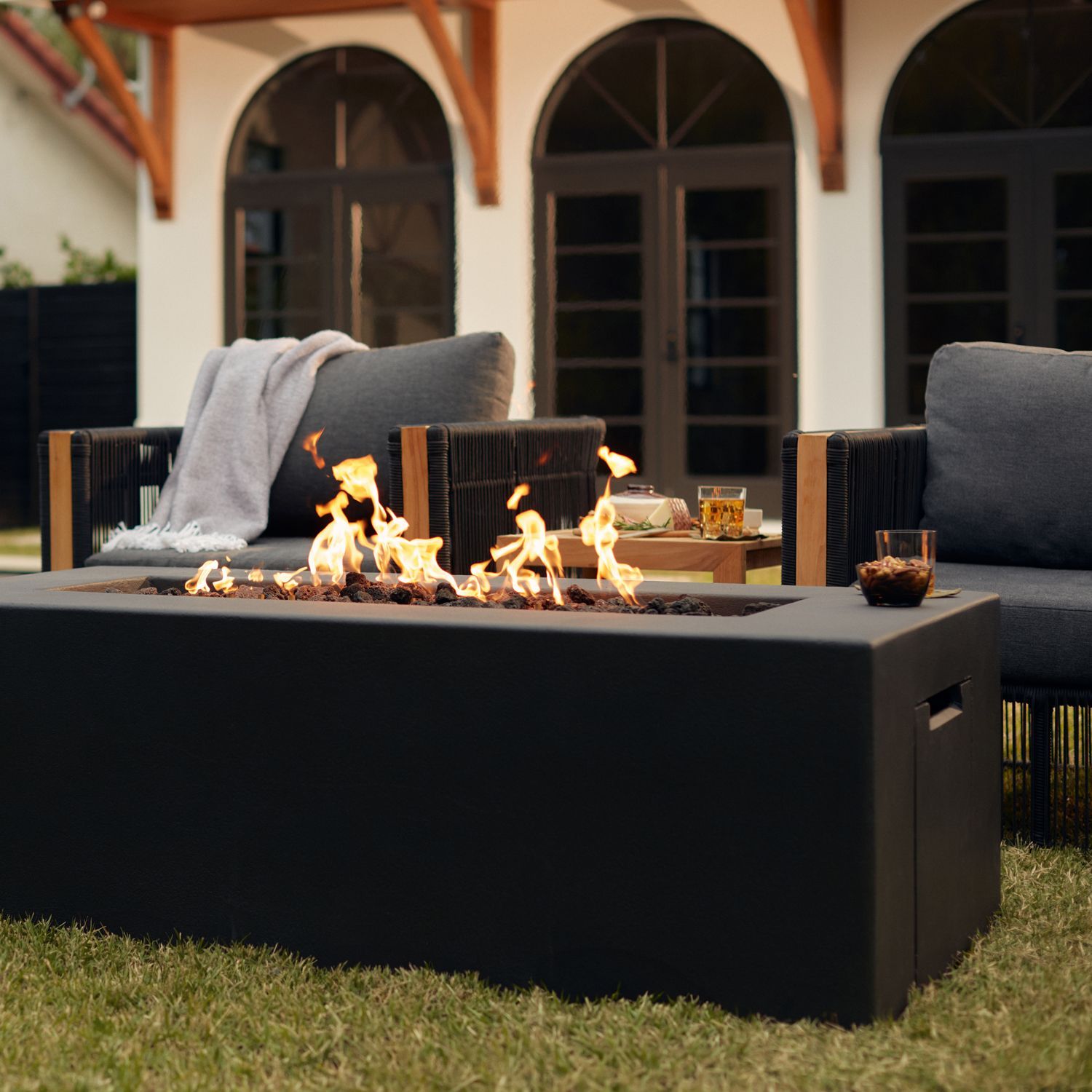 Outdoor patio with a black fire pit table, lit flames, and cushioned seating in front of arched doors