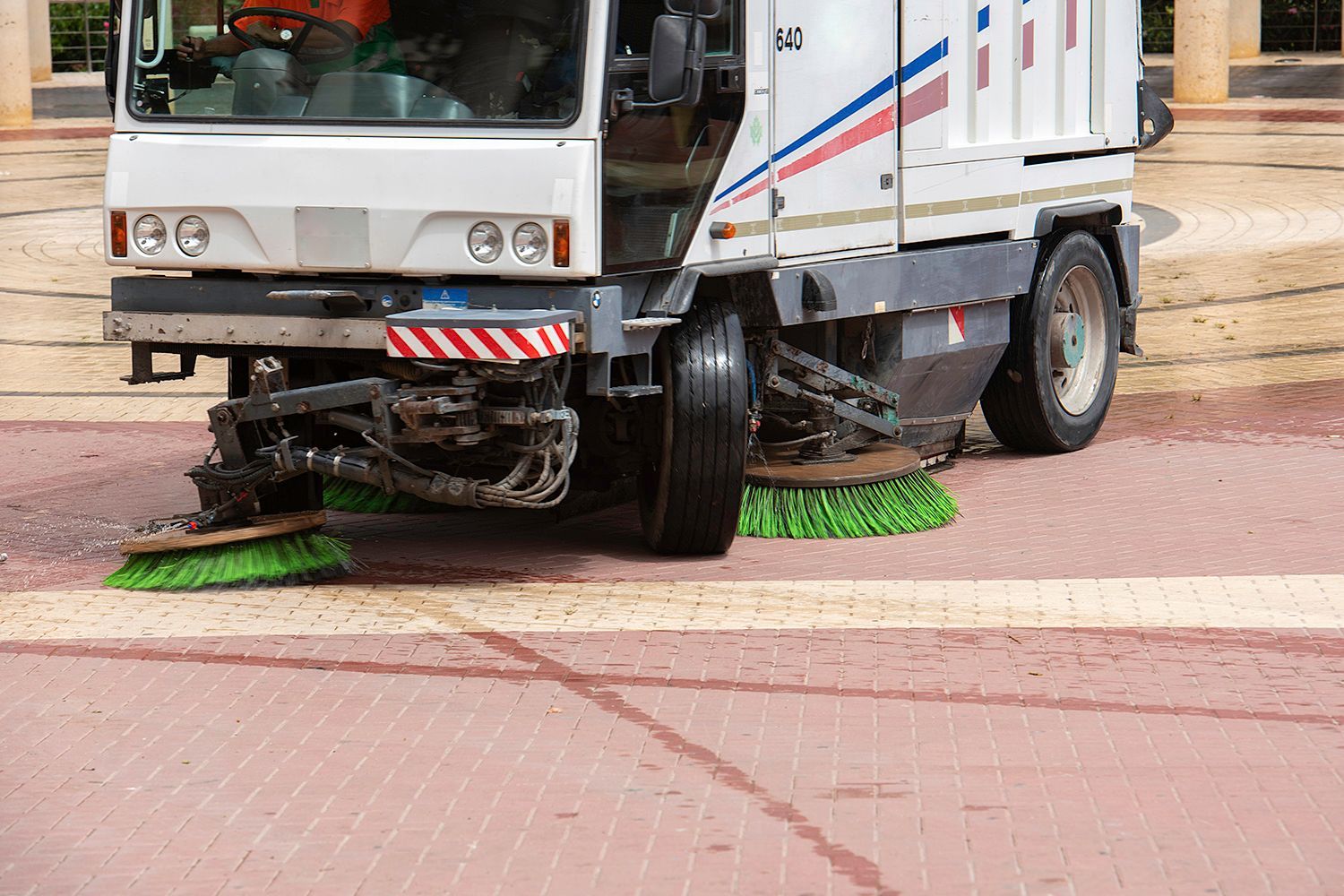 Street sweeper cleaning a red brick paved area. White truck with green brushes in action.