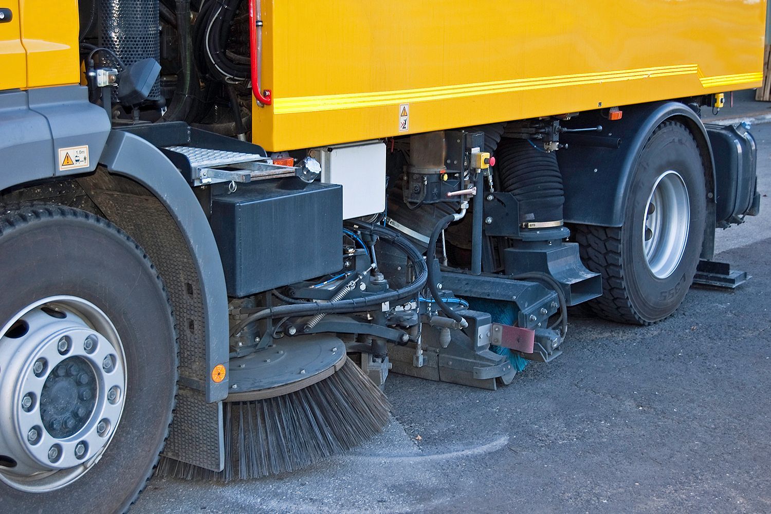 Side view of a yellow street sweeper truck with rotating brushes cleaning a paved road surface.