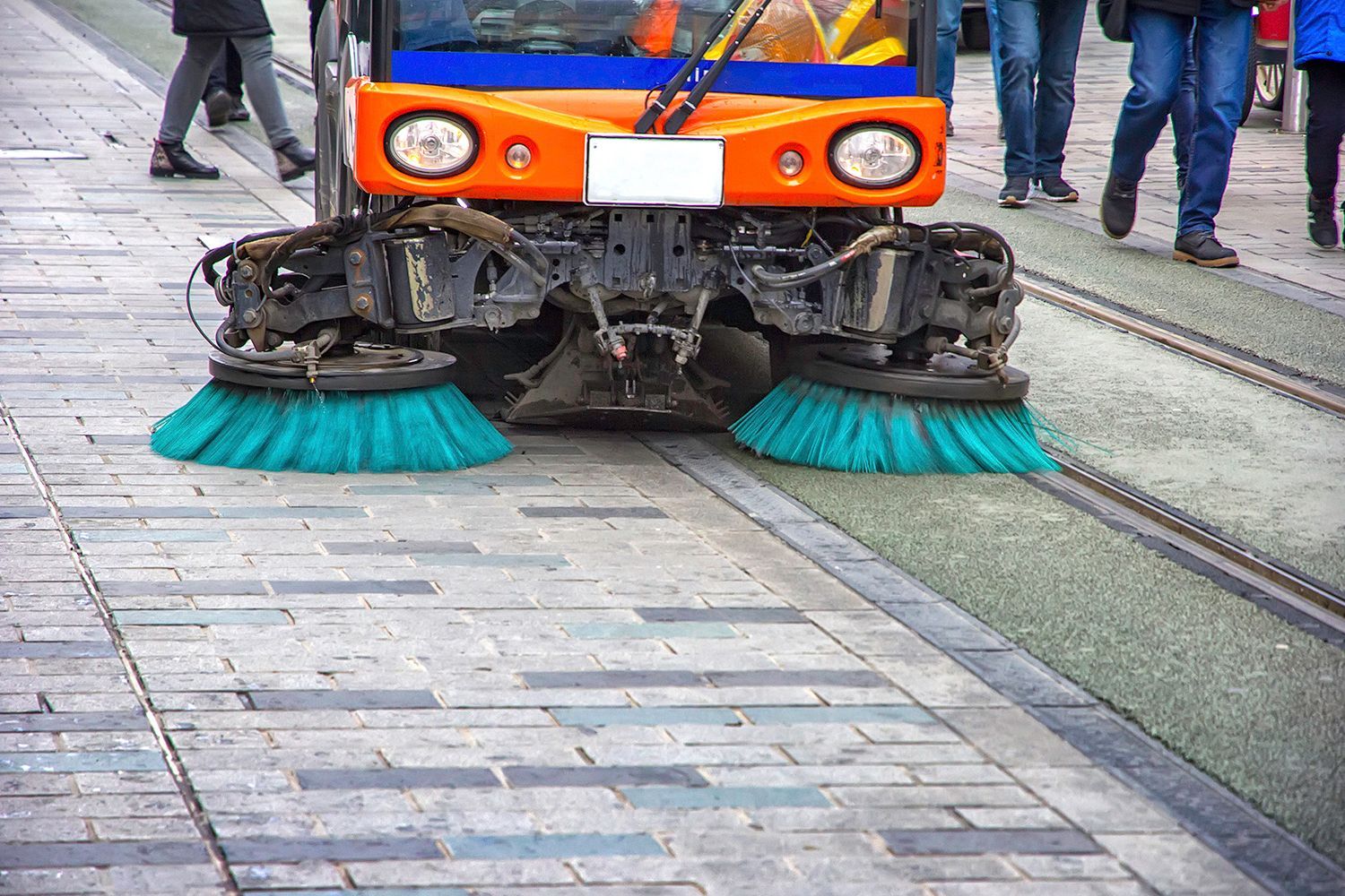 Street sweeper cleaning a brick sidewalk with green brushes, orange vehicle, and people nearby.