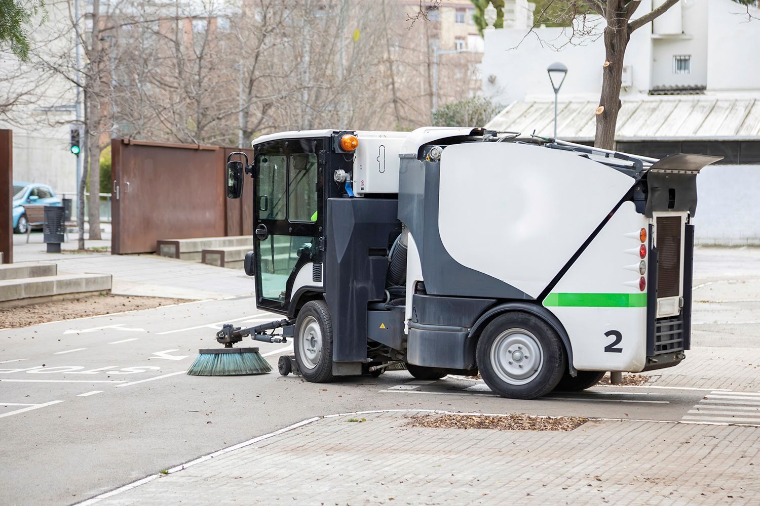 Street sweeper cleaning an asphalt road in a public space. White and gray vehicle, brush sweeping.