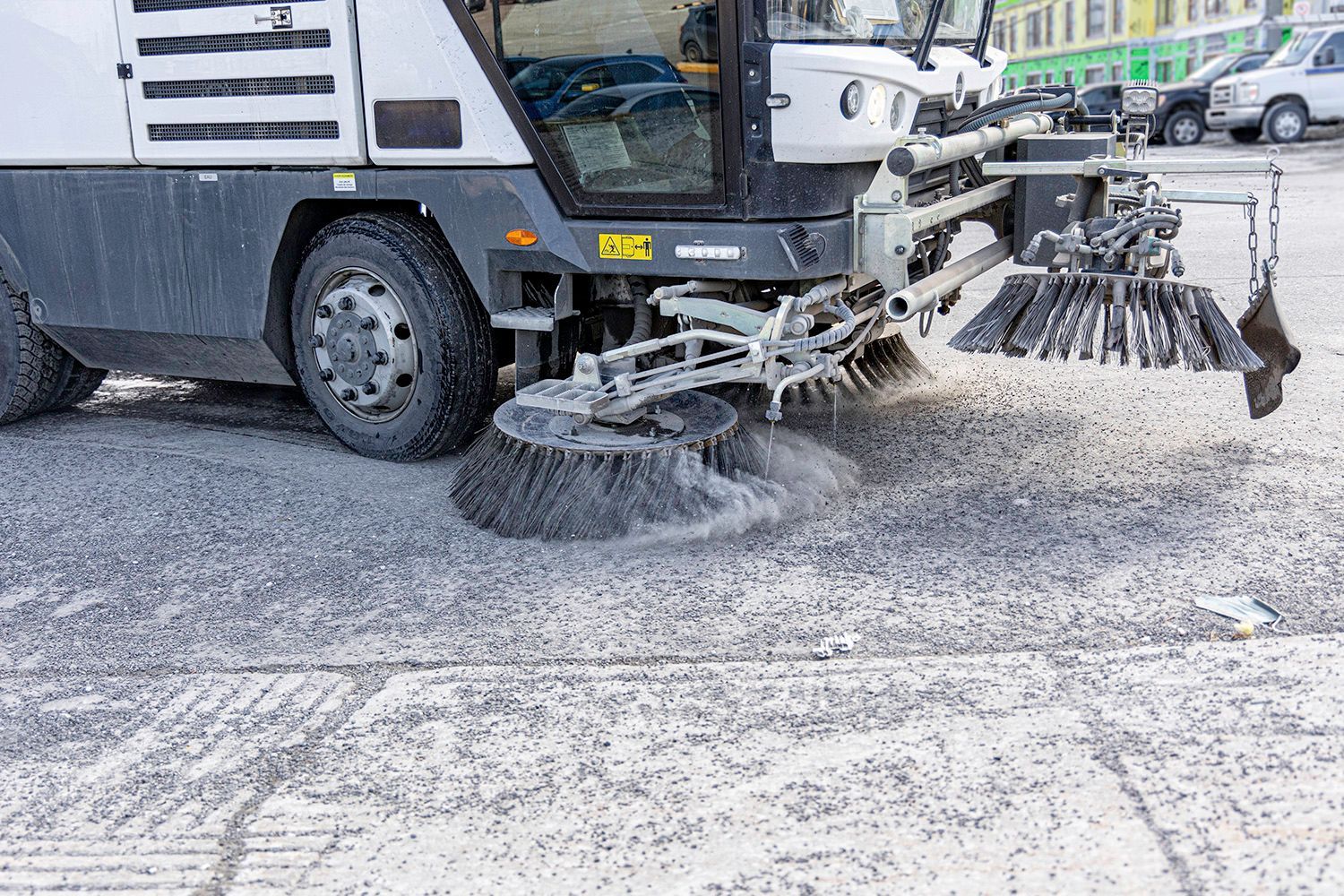 Street sweeper cleaning a snow-covered road with rotating brushes in a city setting.