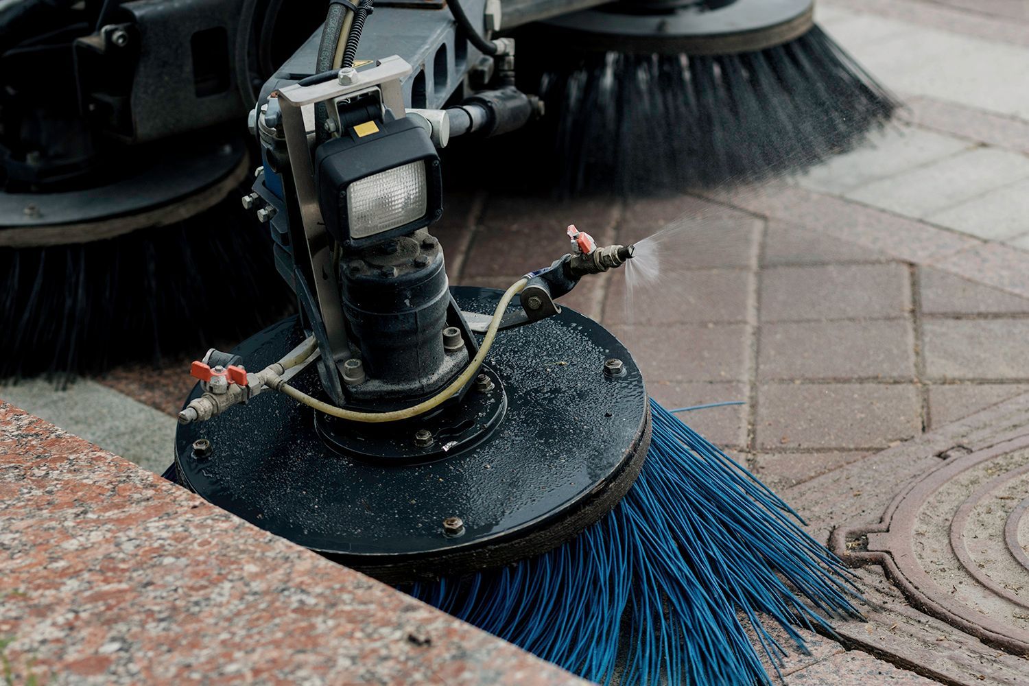 Street sweeper with blue brushes cleaning a tiled sidewalk.