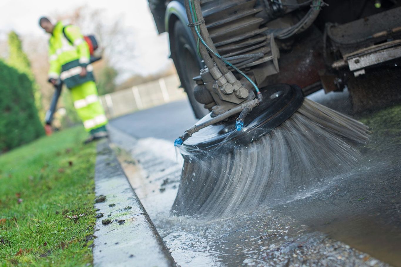 Street sweeper brushes a wet road next to a lawn; worker in reflective gear operates a leaf blower. Street sweeper brushes a wet road next to a lawn; worker in reflective gear operates a leaf blower.