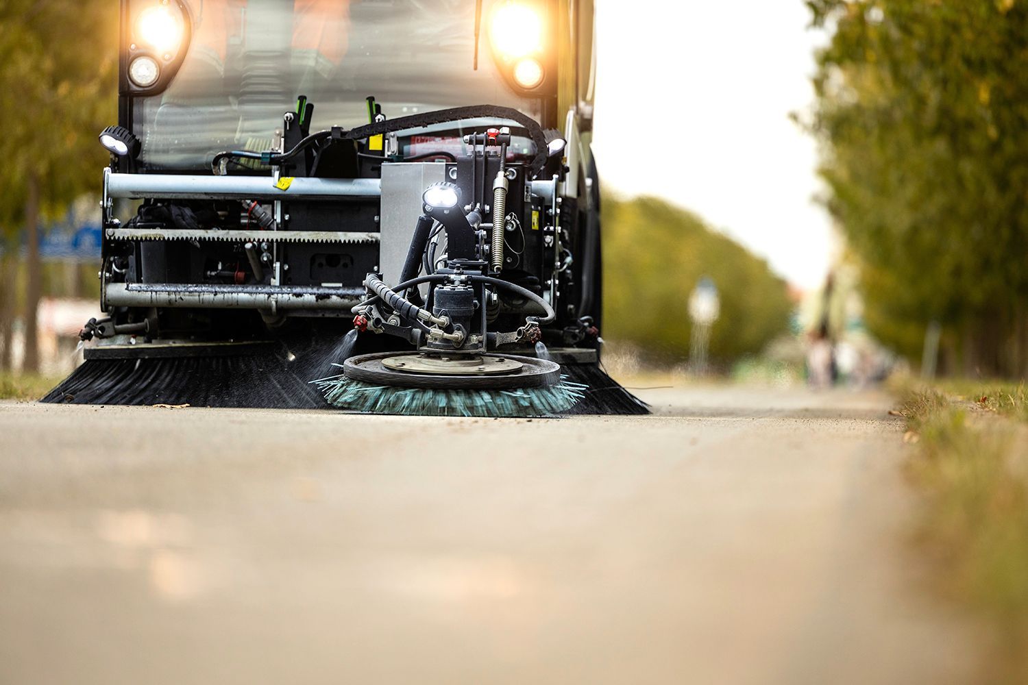 Street sweeper cleaning a road, bristles sweeping debris. Sunlight and trees in the background.