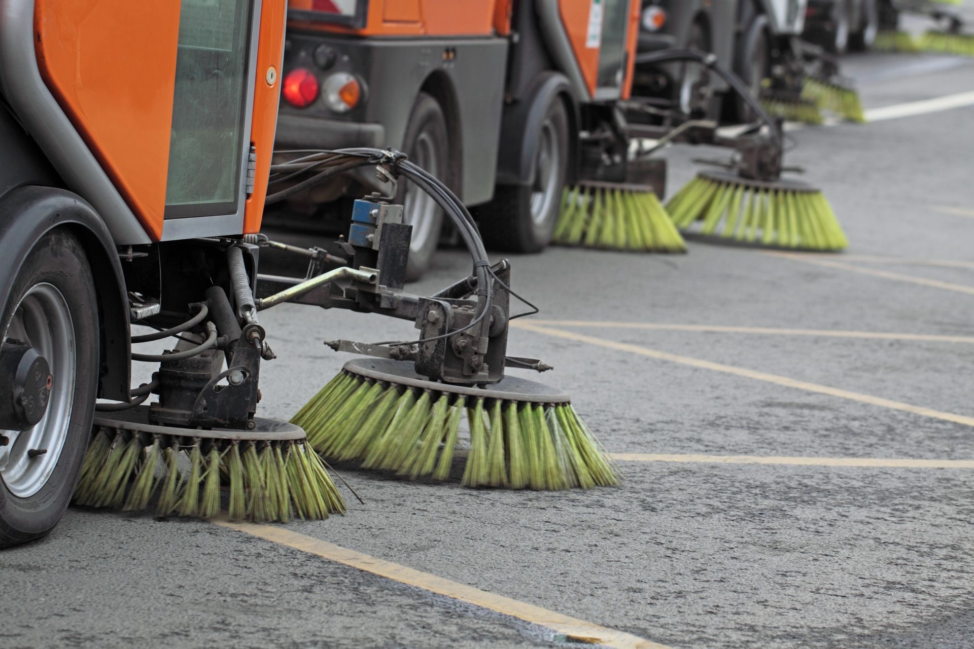 Street sweepers with rotating brushes cleaning a paved roadway or parking lot.