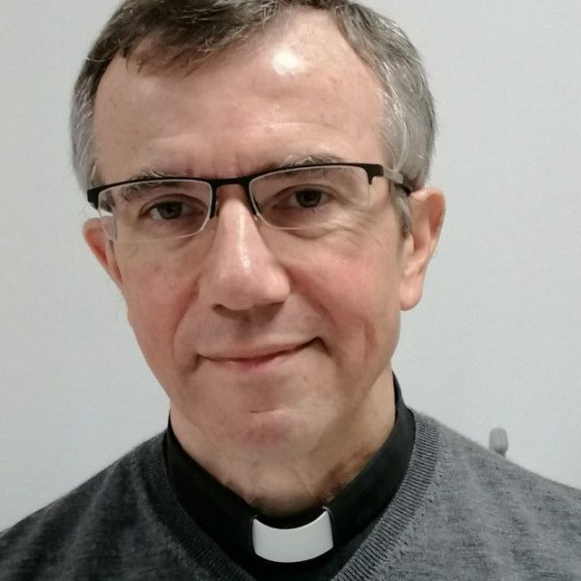 Un uomo con gli occhiali e un collare da clero guarda la telecamera