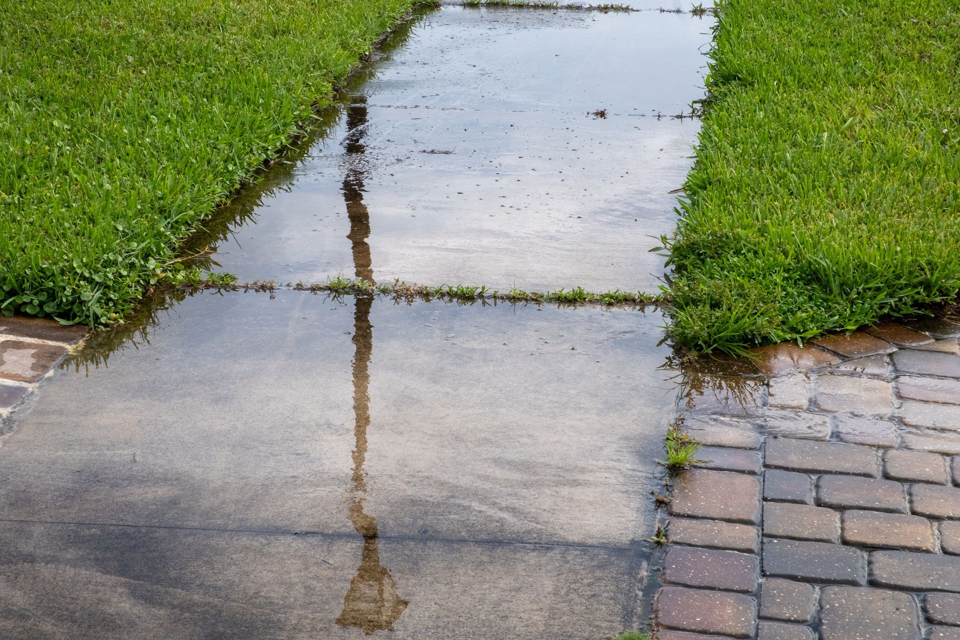 Rain-soaked sidewalk beside green grass and brick paving, with a pole reflection in a puddle