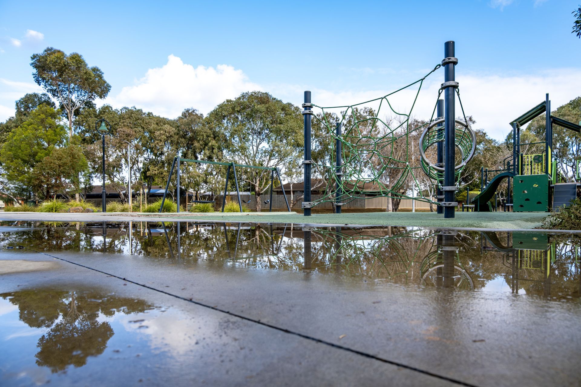 Empty outdoor playground with wet concrete, trees, and a blue sky reflected in puddles