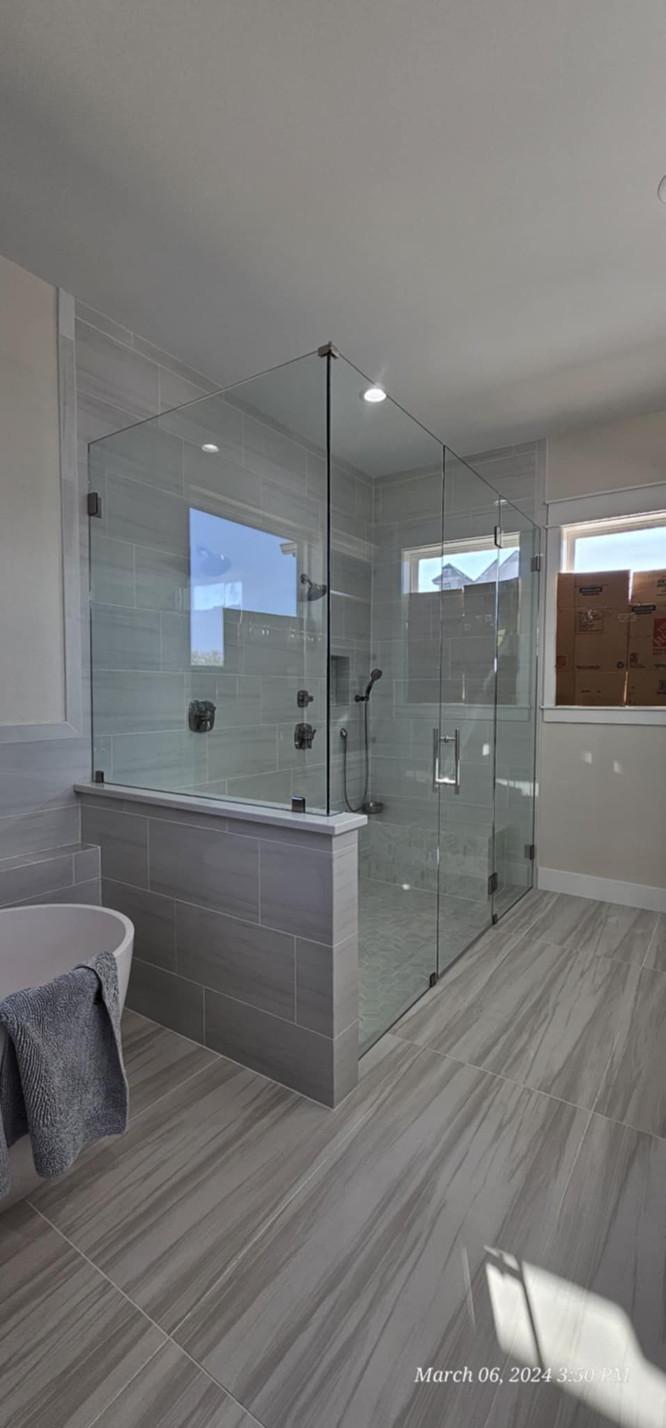 Modern bathroom with glass shower enclosure, gray tiled floor, and a towel on a tub.