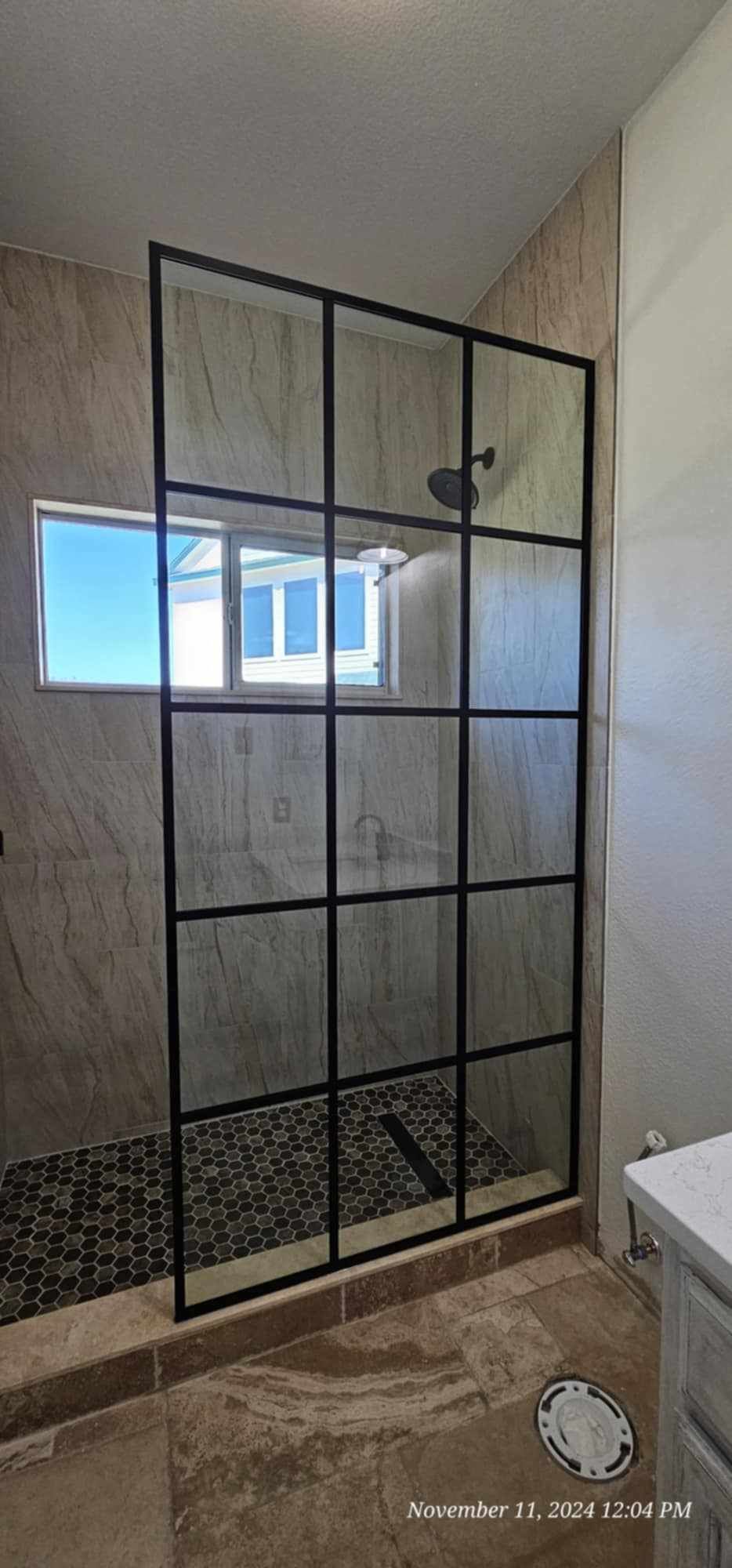 Black framed shower with glass panels; beige tiled shower and floor. Window in the background.