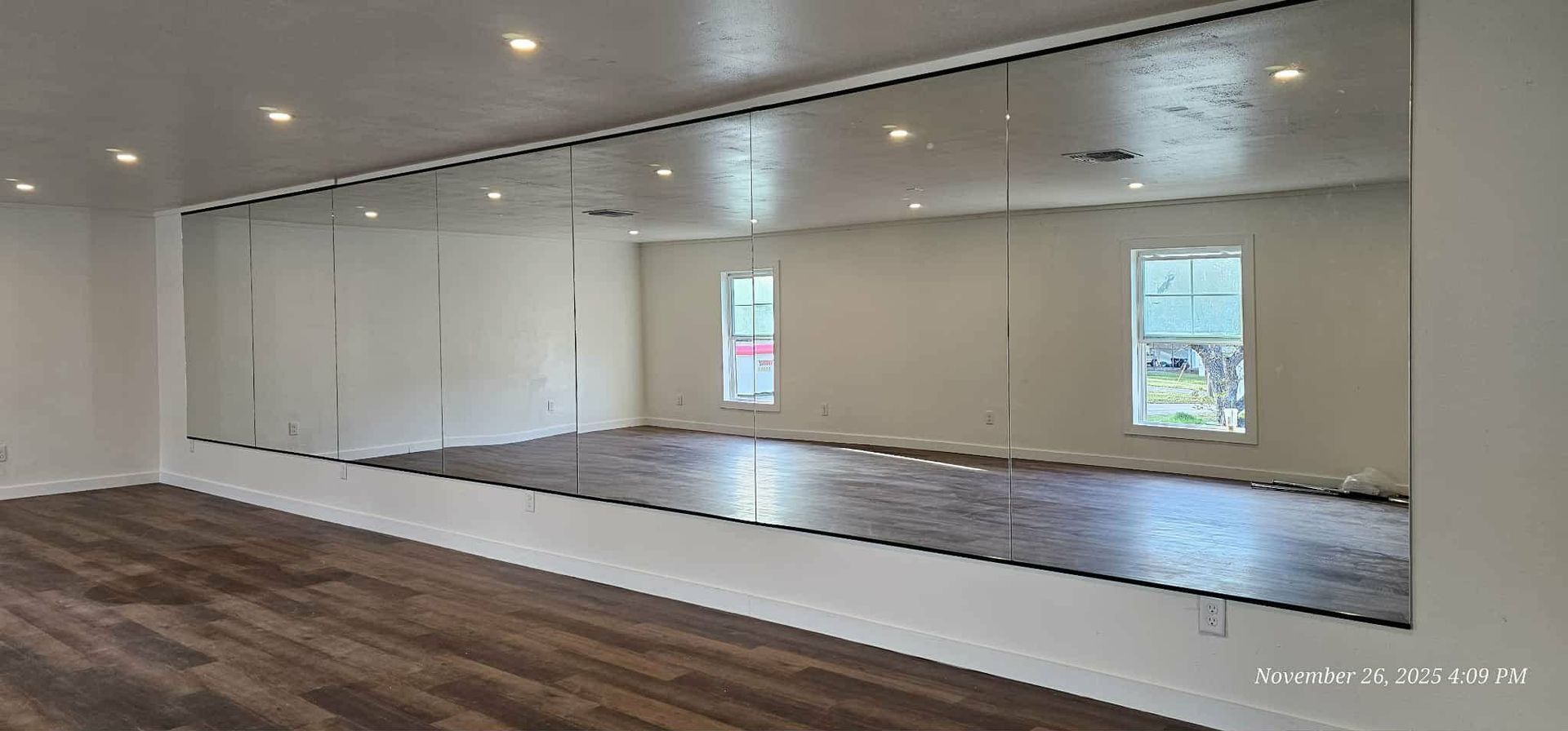Large room with wood flooring, white walls, and a wall-length mirror. Recessed lighting.