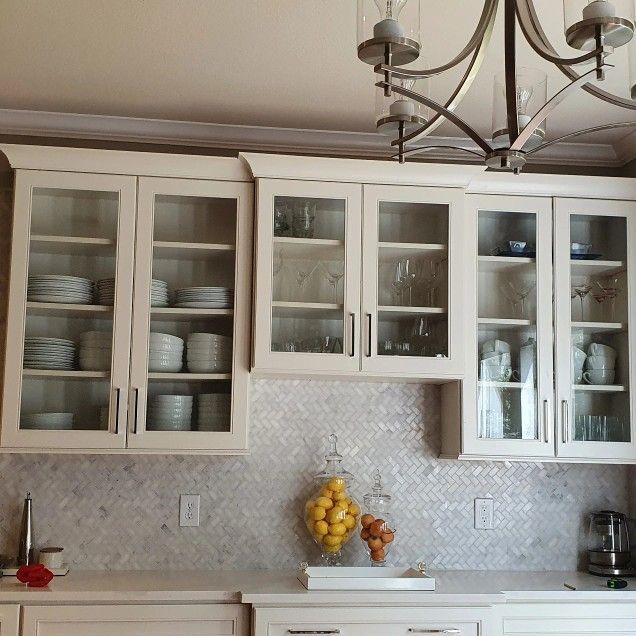 Off-white kitchen cabinets with glass doors, displaying dishware and glassware, above a mosaic backsplash.